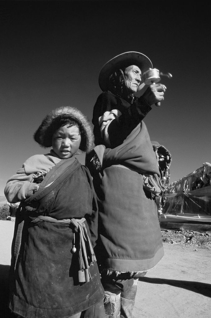 Photo: Black and white photo of two people, one and adult and one a child, wearing traditional rural clothes in China. The adult holds a noise maker in their hands as they look off to the distance as the child turns to look at the camera with an ambivalent look.