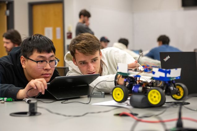 Photo: Yang Yang Zhang (ENG’24) (left), and Kai Imery (ENG’24) work to get their object-detecting autonomous vehicle to run. A young Asian man wearing glasses and a young white man wearing a white hooded sweatshirt both lean close the table in front of them as they study the small electric car they're programming. The small blue car is connected to wires and attached to a small black device that the two are working on.