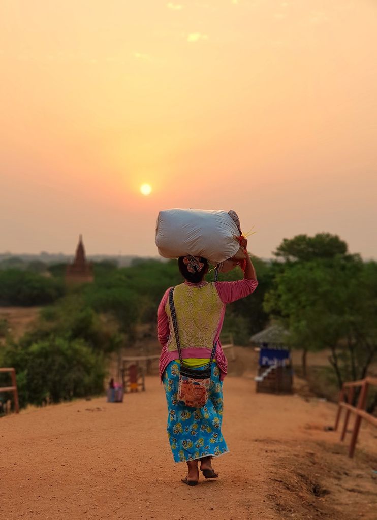 Photo: A long woman walks down a dirt road away from the camera during sunrise in Myanmar. She wears a pink and yellow shirt and bright blue skirt as she carries a large, stuffed bag on her head and balances it with one arm. 