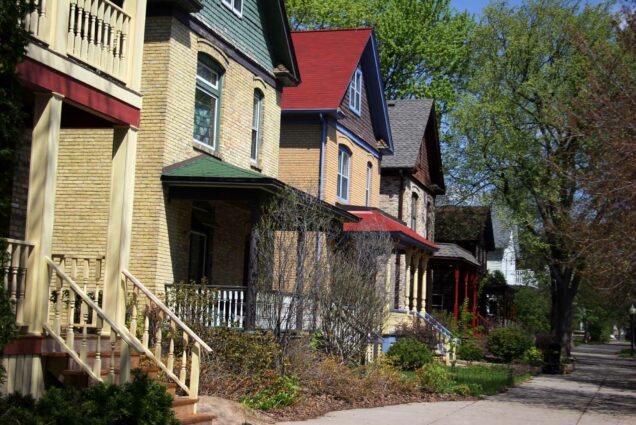Photo: A sidewalk lined with cookie-cutter houses on the left side. They are tan with red, or blue roofs. Most have a porch with an awning on it. There are trees lining the other side of the sidewalk. The day is bright and sunny.