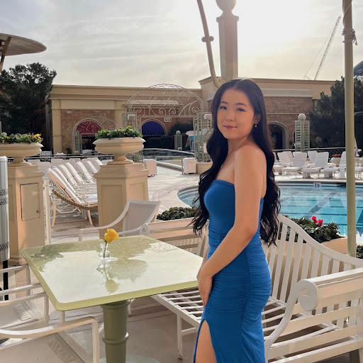 This is BU student, Irene Kim, and she is standing with a blue dress on, smiling at the camera. She is outside at a hotel, in front of a table and chair set by a pool. 