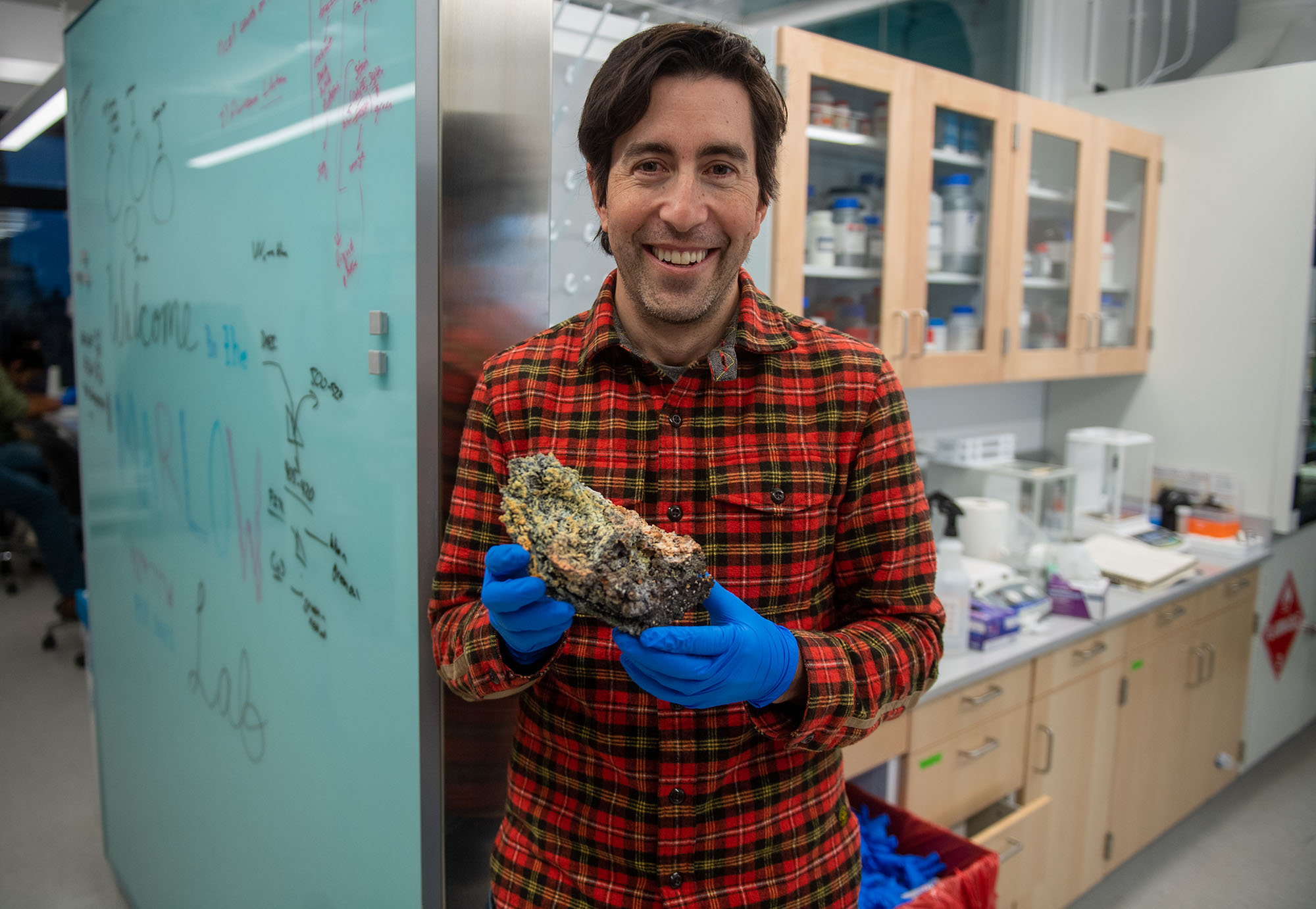Photo:  Jeffrey Marlow, a white man with dark brown hair and wearing a red, black and yellow plaid shirt and blue medical gloves holds up a volcanic rock as he poses in his lab.
