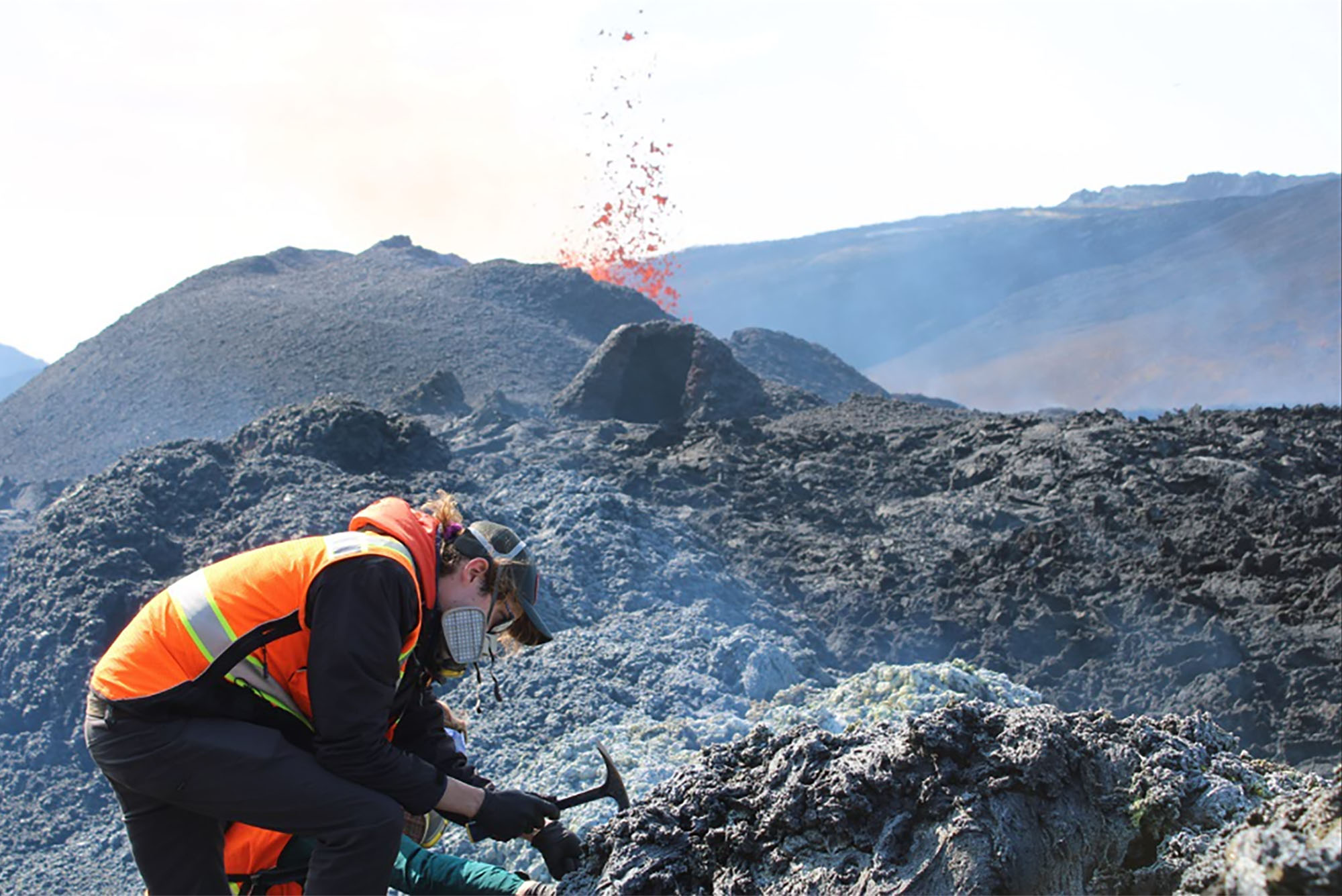 Photo: Iceland’s Fagradalsfjall volcano erupts in the background as Peter Schroedl, a person wearing black clothes, a face mask, orange neon vest collects specimens from volcanic rocks in the foreground.