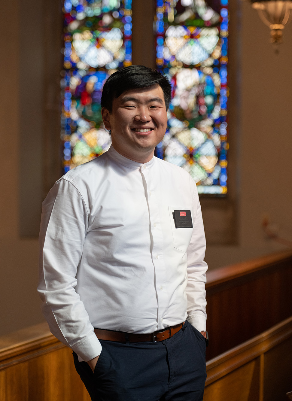 Photo: Jonathan Lee, an Asian man wearing a white long-sleeved chaplain shirt, black pants, and a silver name tag that reads "Jonathan Lee" sits posed in a pew. He smiles, hands in pockets as he looks to the camera in a church.