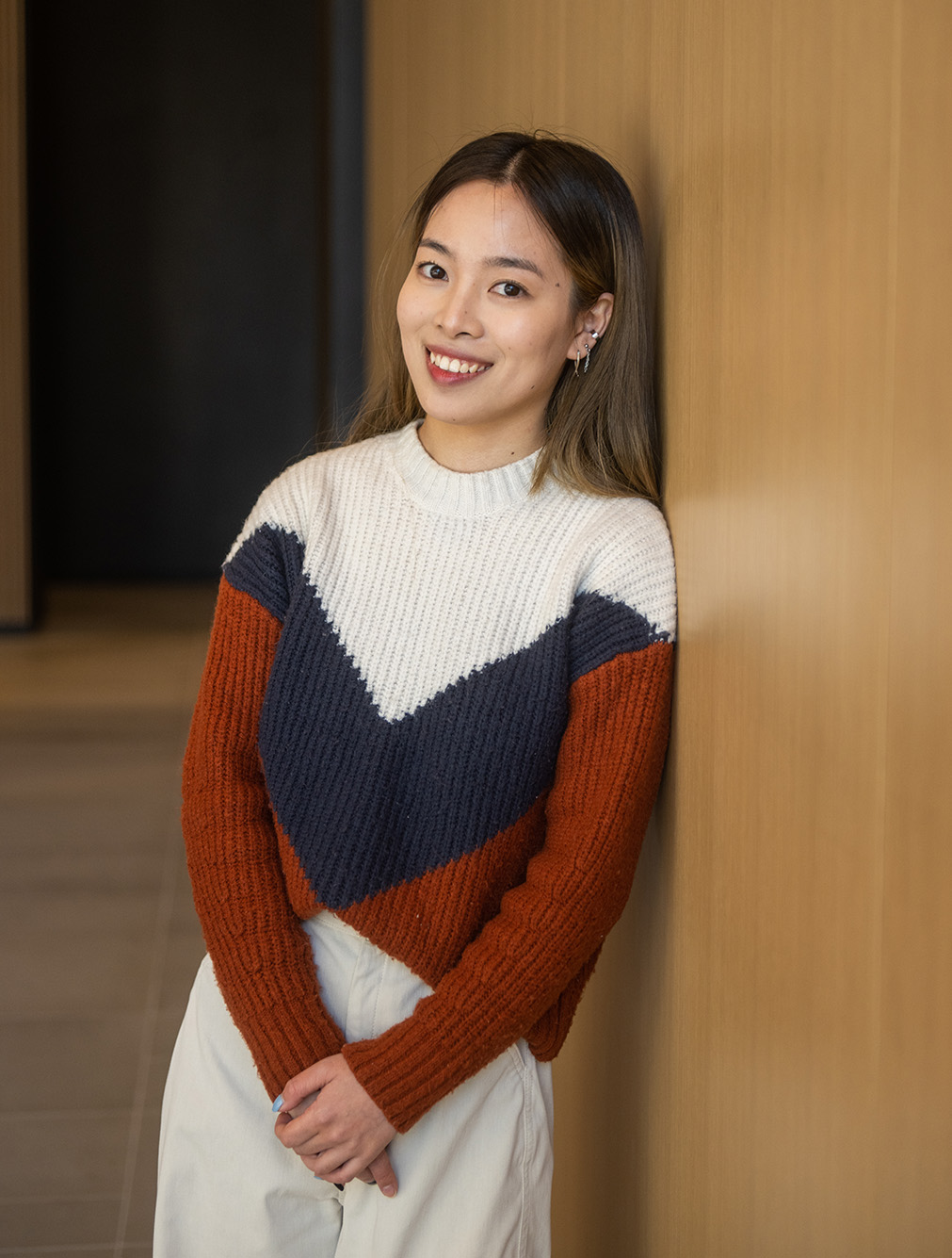 Photo: Linzi Scholarship recipient Liyu Chen poses against a wooden wall. A young Asian woman wearing a white, red, and black sweater, and long white skirt, smiles as she poses next to a light wooden wall.