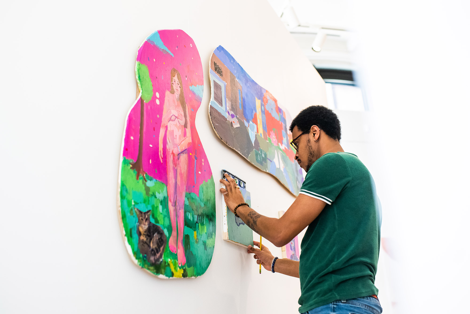Photo: A Black man adjusts a small painting on the wall for his MFA thesis exhibition. The painting on his left is a curvy canvas (looks like an outline of the number 8) with a pink background and a nude woman on the front. The top painting is of a landscape with houses in the same form as the one on the left.