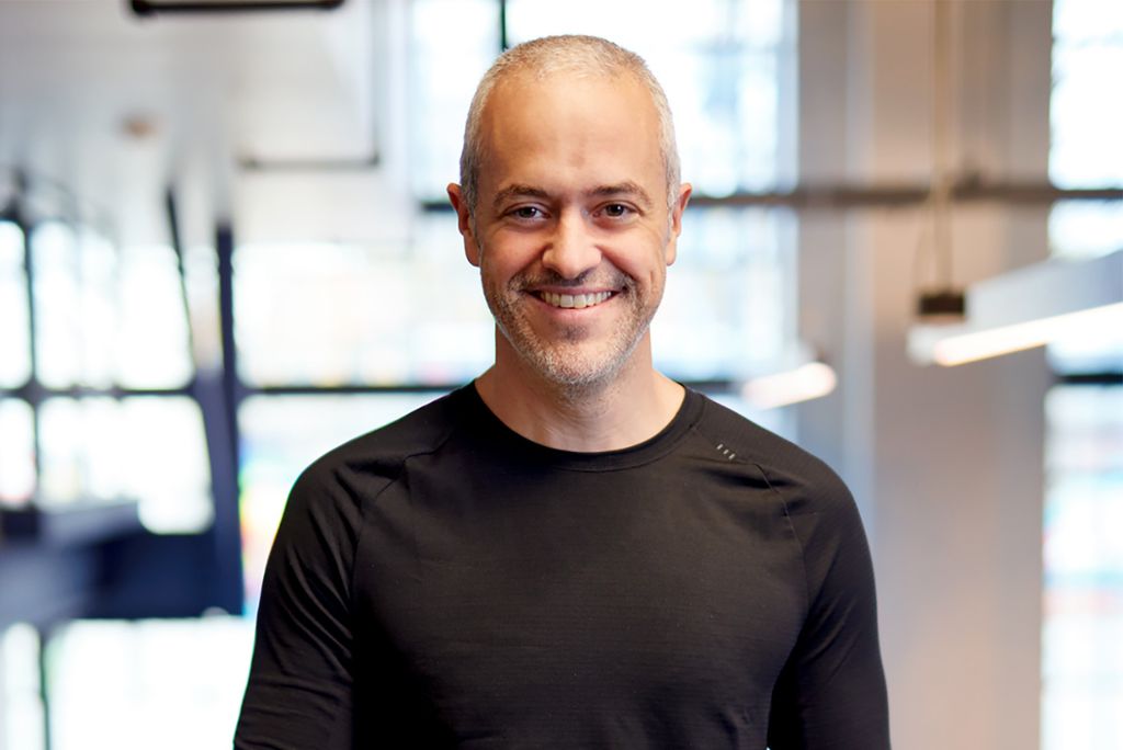 Photo: Mike Salguero, a white man with short, buzzcut silver hair and wearing a long sleeved black shirt, smiles and poses for a photo. The blurry background is brightly lit.