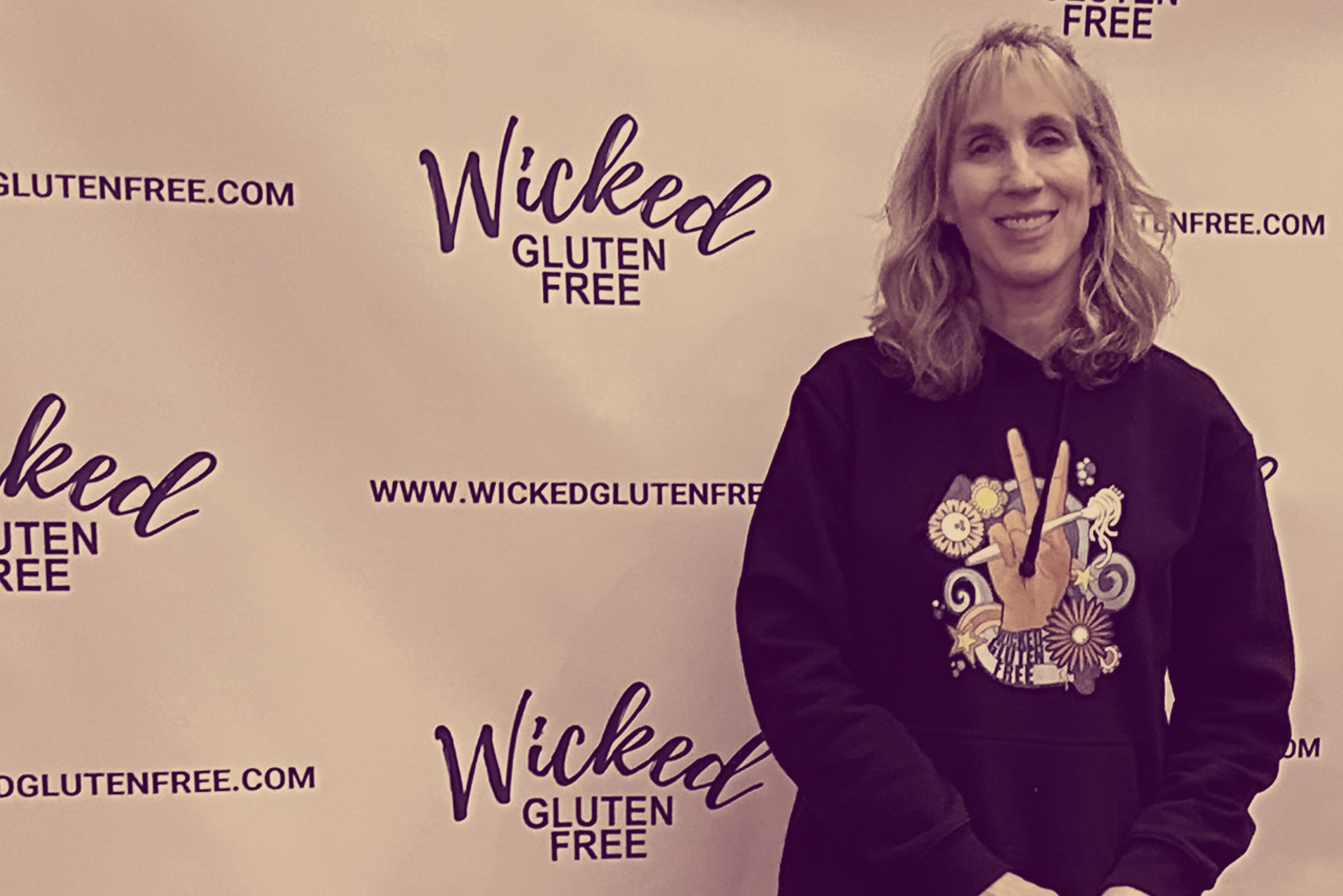 Photo: Abby Helman Kelly, a white woman with short, wavy blonder hair and wearing a black hooded sweatshirt and jeans, poses in front of a white backdrop that features a logo that reads "Wicked Gluten Free" poses with a sign in her hands that she leans on. Sign reads "Wicked Gluten Free". Image has a color overlay that tints the photo a burnt yellow.