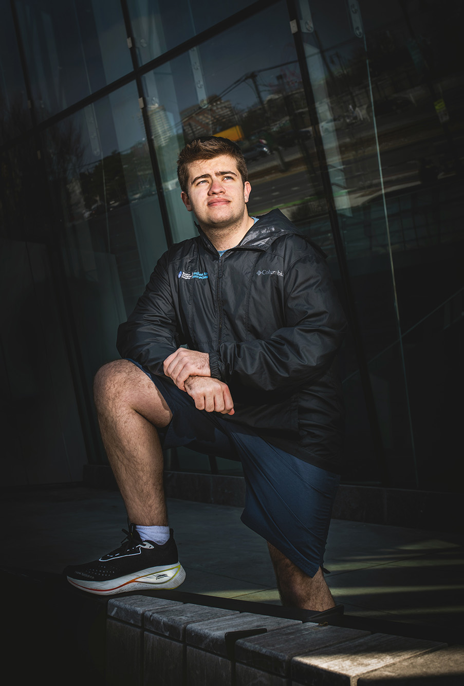 Photo: Nico Iannacone poses for a photo. A white young man wearing a black rain jacket and black shorts poses with one leg resting up against a wall as he looks of to the distant. Photo has a moody theme to it.
