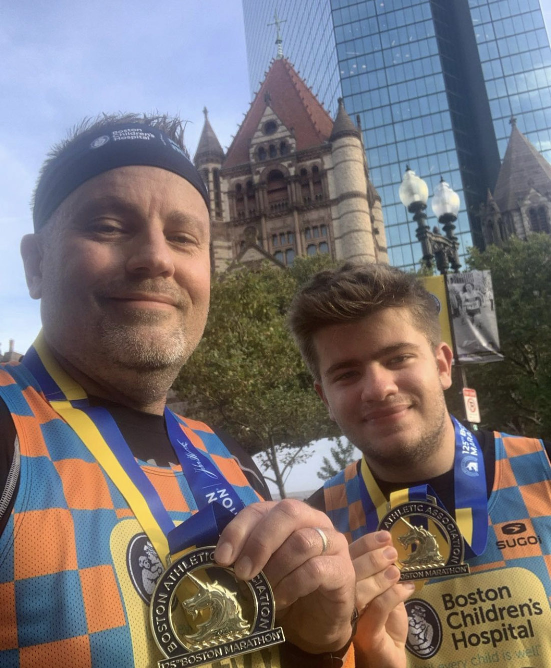 Photo: Two men wearing identical orange and blue checkered shirts take a selfie together as they pose and hold up golden Boston Marathon medals.