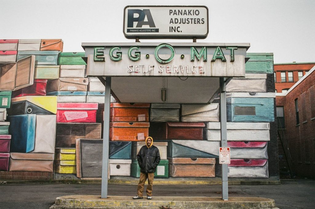 Photo: A man wearing a yellow hood, leather jacket, and tan pants stands in underneath a large overhang that reads "Egg-O-Mat Self-Service". A large mural featuring a variety of shoe boxes covers the large brick wall behind him as he looks forward at the camera.