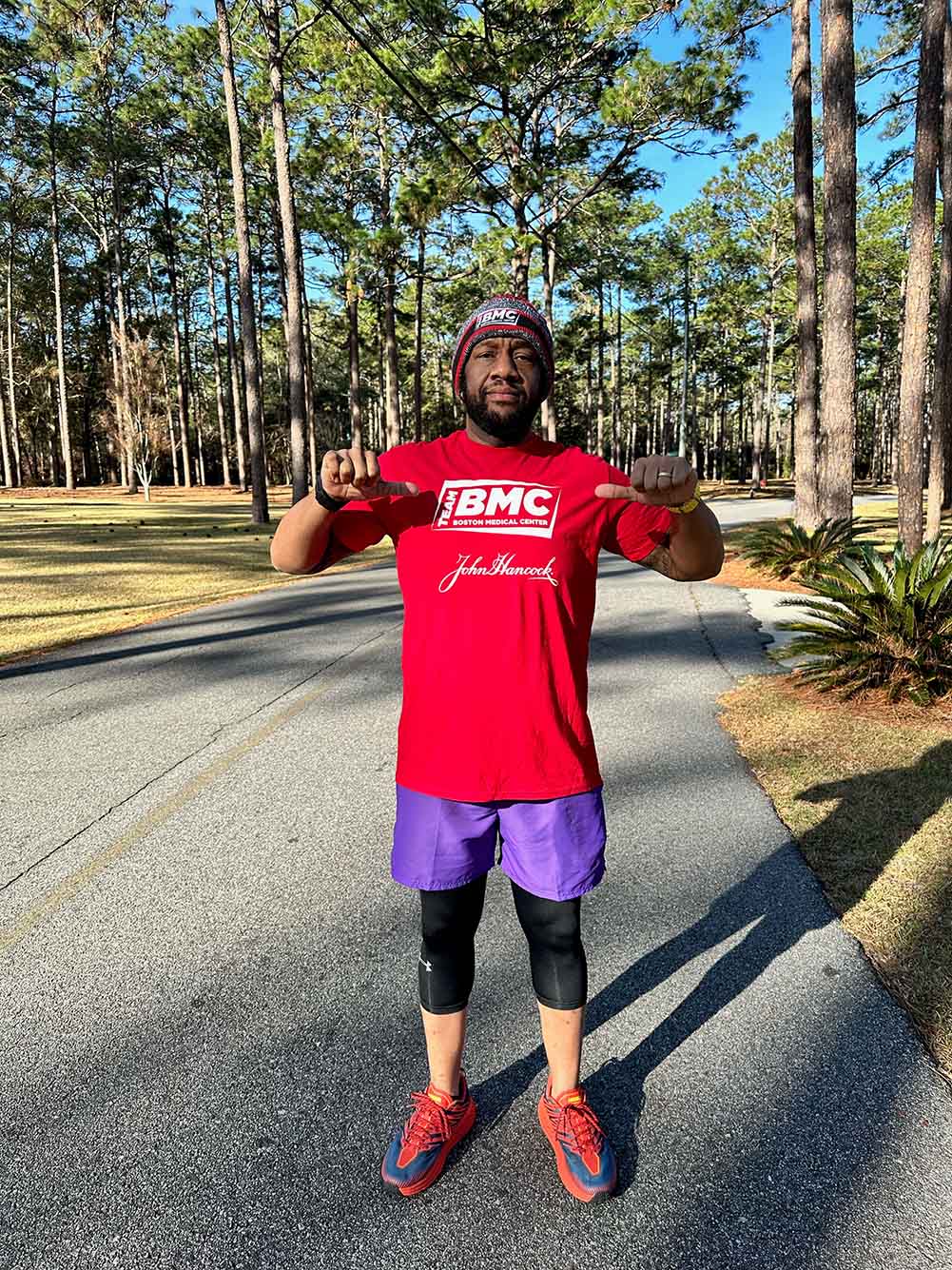 Photo: Frederick Powell, a black man wearing a red short-sleeved shirt that reads "BMC" and purple running shorts, poses on a running trail in woody area.