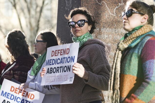 Photo: Lindsay London, a white woman with short brown hair, holds protest sign in front of federal court building. She stands with other protestors in support of access to abortion medication. The protestors are bundled up in scarves and fleeces. They have their sunglasses on and they stand in front of a tree.