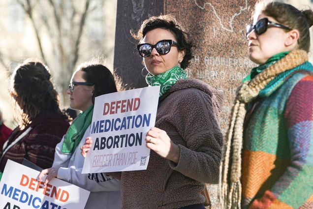 Photo: Lindsay London, a white woman with short brown hair, holds protest sign in front of federal court building. She stands with other protestors in support of access to abortion medication. The protestors are bundled up in scarves and fleeces. They have their sunglasses on and they stand in front of a tree.