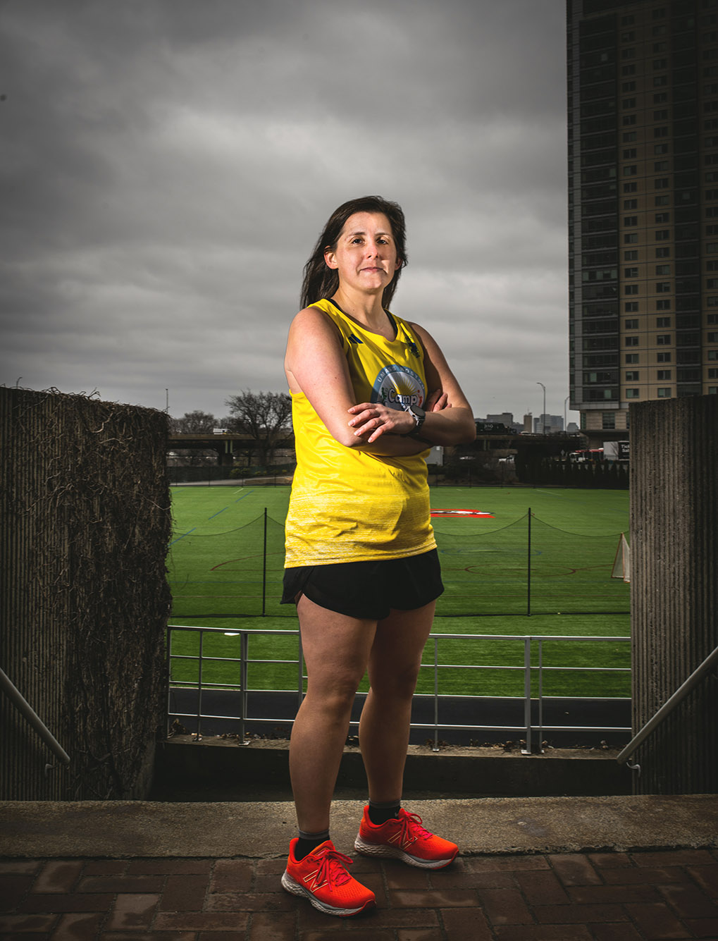 Photo: Sara Tenenholtz, a white woman wearing a yellow Boston Marathon running jersey and black running shorts poses with arms crossed in front of a Nickerson soccer field for a moody photo.
