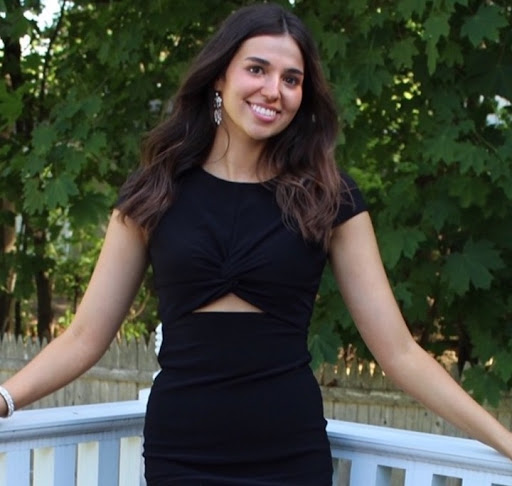 This picture is of a girl and she is outside with a black dress on. She is a deck and a green tree is behind her. She has her hands on the railing and is smiling to pose for the picture. 