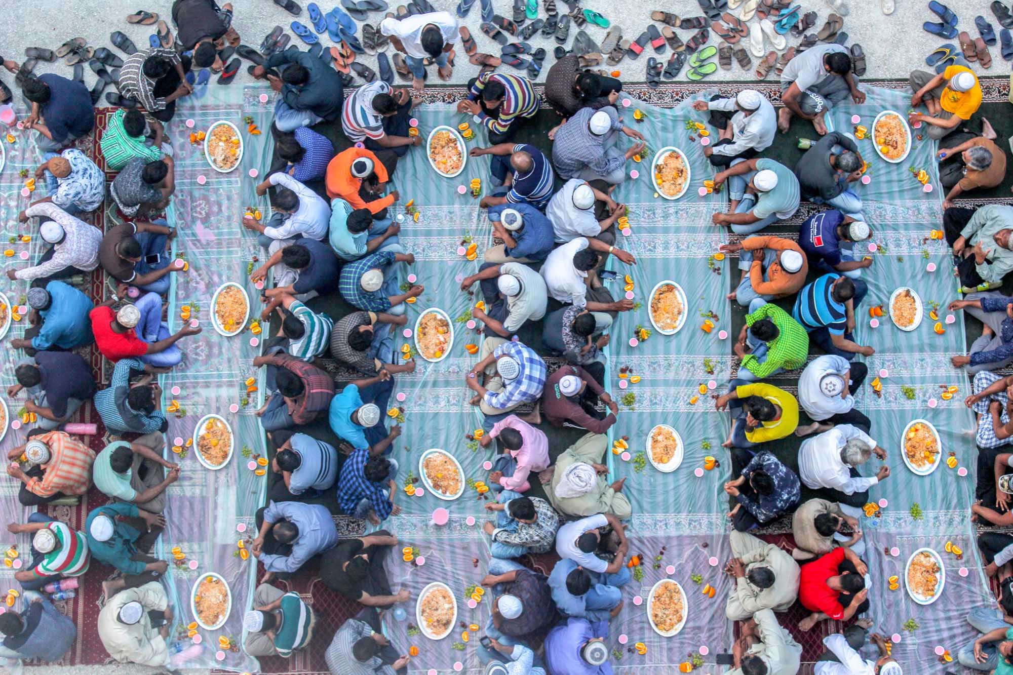 Photo: Overhead shot of rows of people sharing and eating food as they sit on a large, colorful cloth outside.