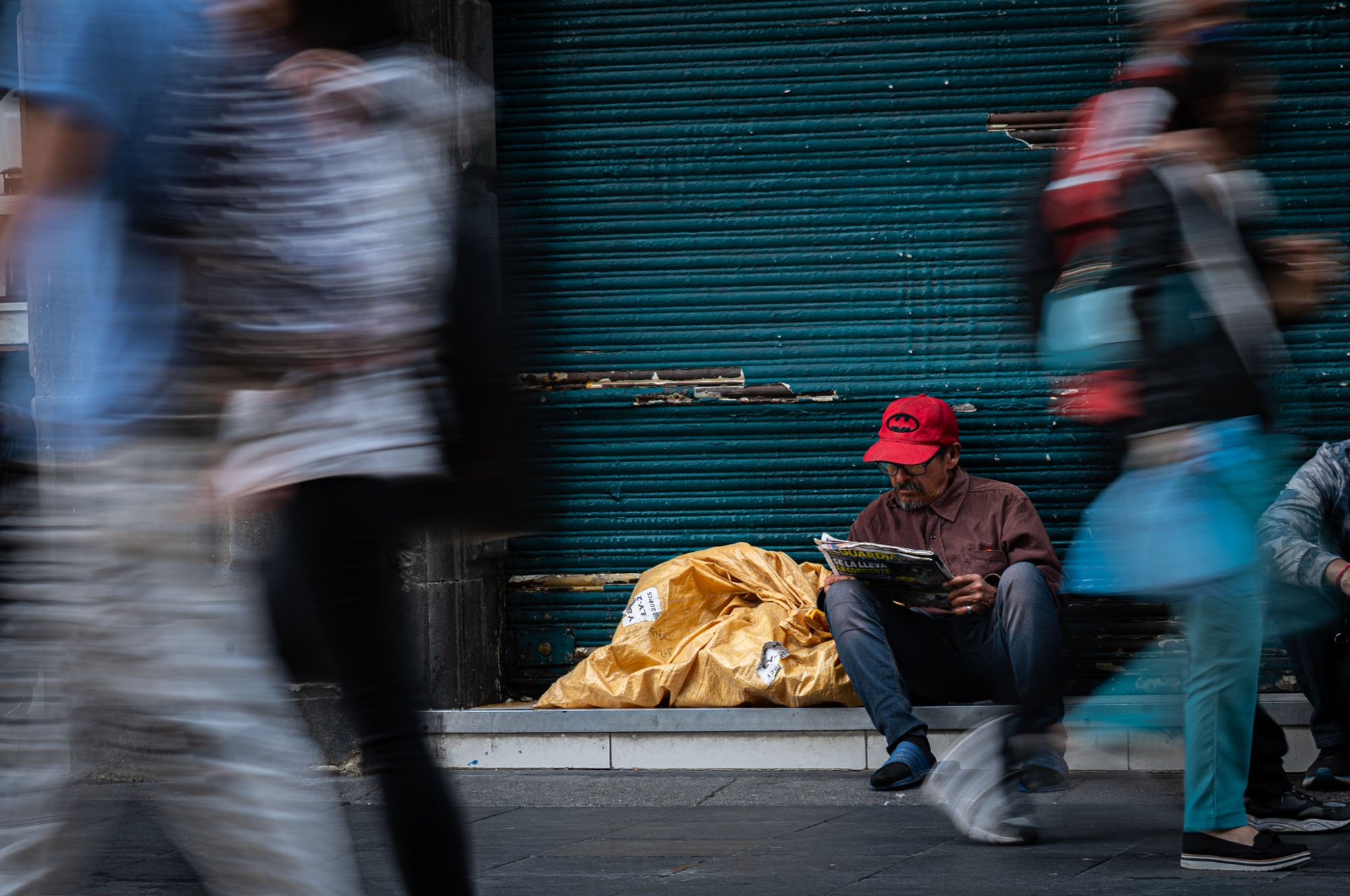 Photo: On a bustling shopping street in the tourist hotspot that is Centro Historico in CDMX, an elderly man wearing a red baseball cap sits by the sidewalk, reading a newspaper, seemingly unaffected by the blurry pedestrians walking by.