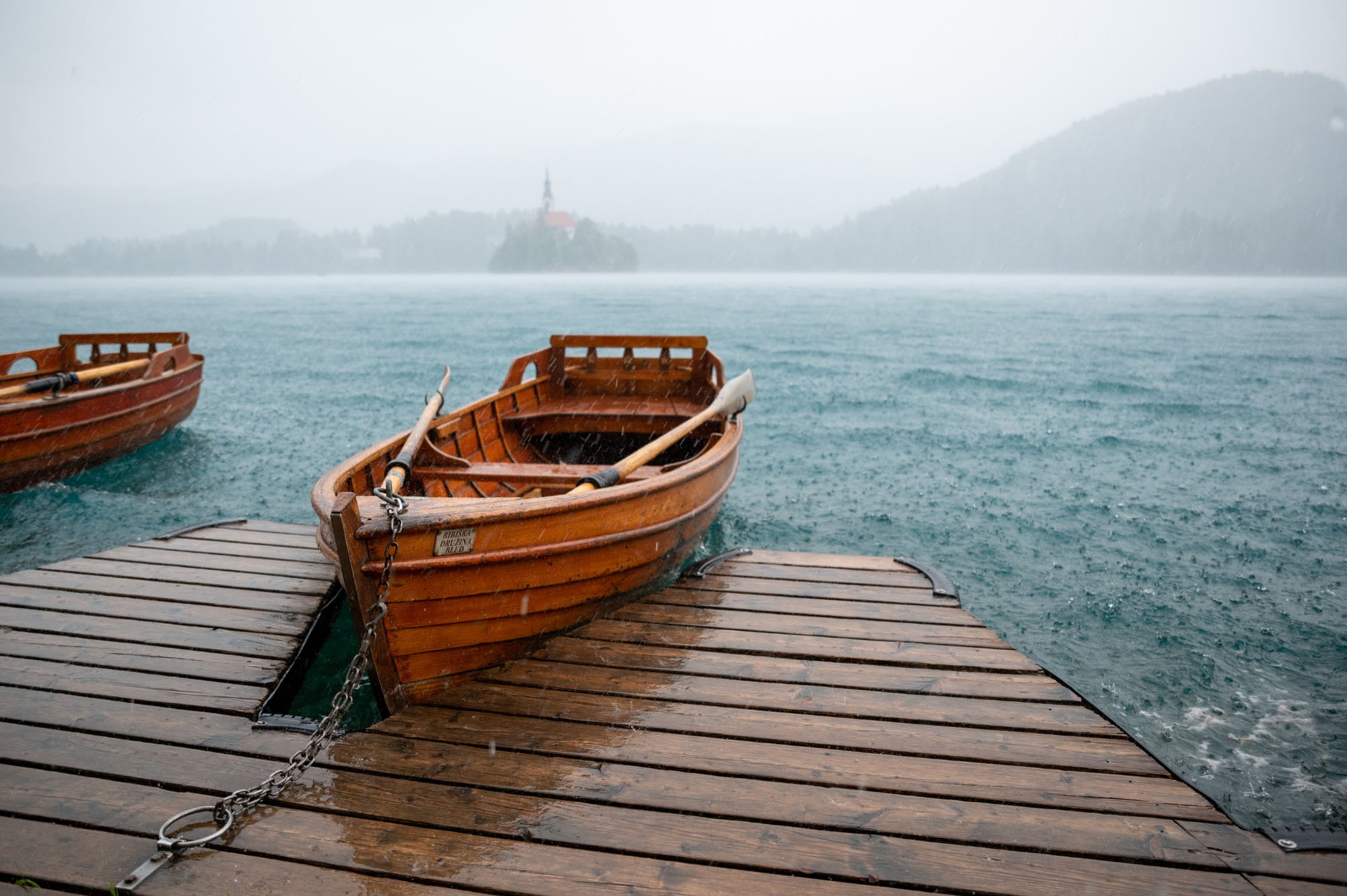 Photo: The Slovenian Olympic rowing team boat is shown docked at a wooden dock as it rains heavily at Lake Bled. The raindrops can be seen dramatically hitting the water. In the distance, a large boar is seen heading towards the dock.