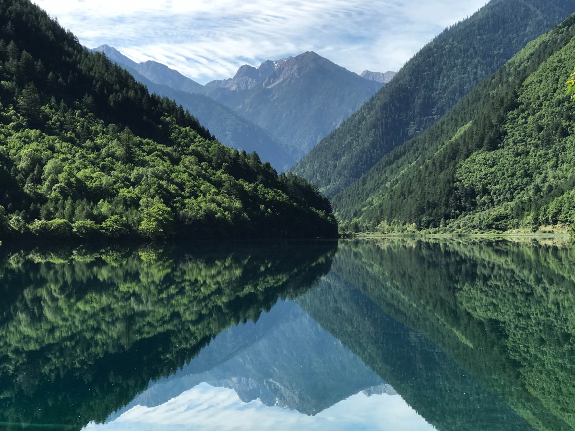 Photo: A large lake is shown surrounded by mountains and large hills filled with lush, green trees. The lake reflects the trees and  scenic clouds in the sky.