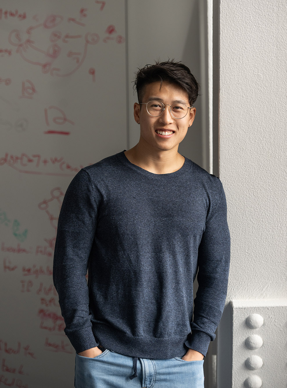 Photo: Lu Lingzi scholarship recipient Zilu “Pete” Tang. A young Asian man wearing glasses and a long sleeved blue-gray shirt, smiles and poses next to a cement wall. Behind him, a white board filled with notes on equations can be seen.