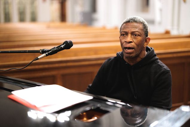 Photo: The choir director of Inner Strength Gospel Choir, Herbert Jones, leads his choir by demonstrating what to sing. His mouth is agape in the photo, mid-singing, and he wears a black hoodie.