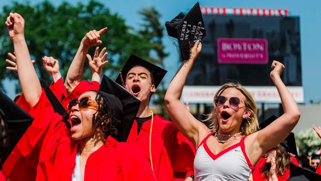 This is photo taken at a past Boston University commencement ceremony. It is outdoors on a sunny day, with students in their caps and gowns shown, cheering with their arms up. Photo Credits: Janice Checchio