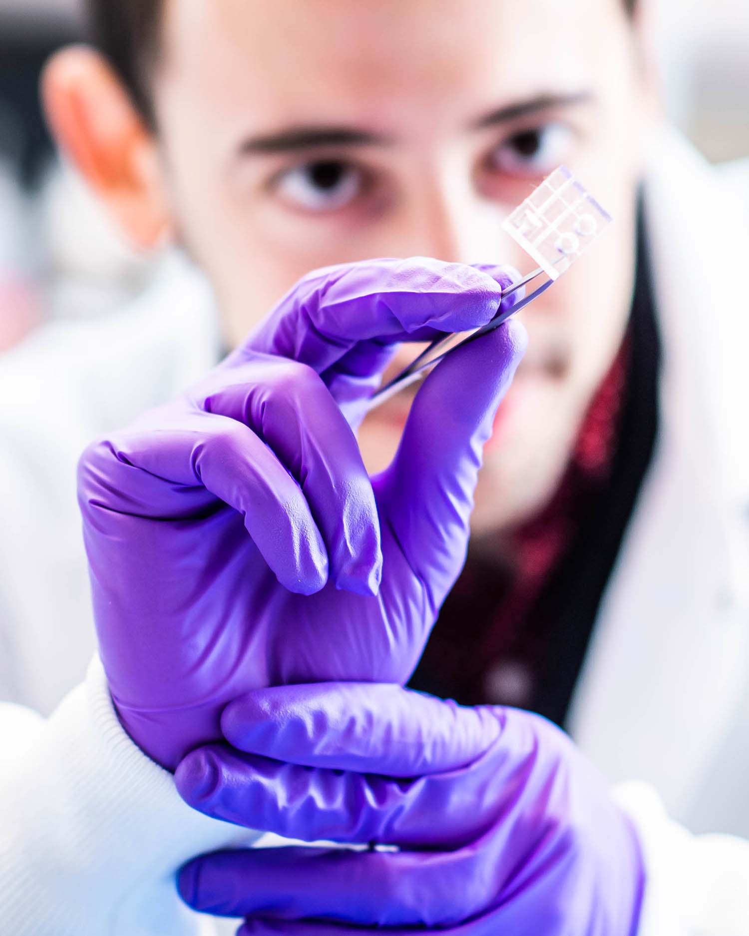 Photo of Christos Michas, a white man with short brown hair, wears purple medical gloves and holds a miniaturized cardiac ventricular pump (or, a mini heart pump) with a pair of tweezers. The pump looks like a small piece of plastic. Michas is seen blurred in the background and the pump and his hands are in focus.