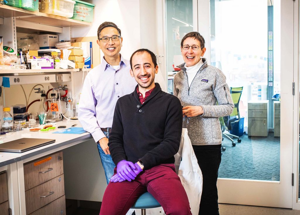 Photo of Dr. Chris Chen (left) postdoc (and former PhD student) Christos Michas(center), Dr. Alice White in their lab on March 7, 2022. Chen is Asian middle aged wearing a gray button-down shirt and glasses. Christos wears purple gloves, a dark gray sweater, and crimson pants and smiles. White wears a gray patagonia quarter zip, black pants and glasses. Behind them, a shelf of lab equipment is seen. 