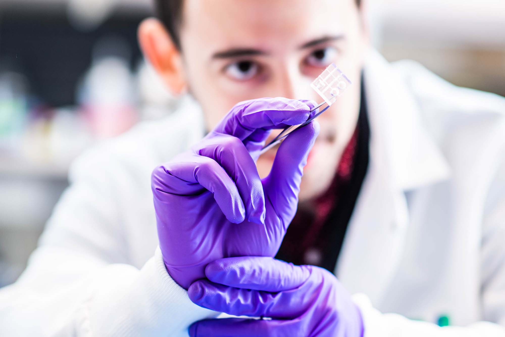 Photo of Christos Michas, a white man with short brown hair, wears purple medical gloves and holds a miniaturized cardiac ventricular pump (or, a mini heart pump) with a pair of tweezers. The pump looks like a small piece of plastic. Michas is seen blurred in the background and the pump and his hands are in focus.