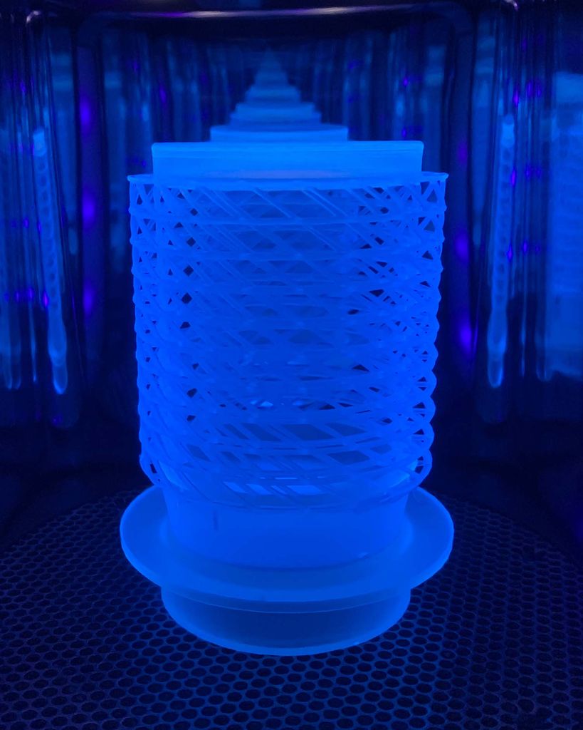 Photo of a large scale replica of the scaffold that supports the heart tissue. The replica is about the size of a computer mouse and looks like a cylindrical plastic strainer. It appears with other replicas on either side in a small container lit with a blue light.