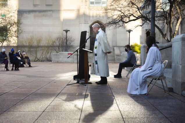 Photo: A horizontal shot of a priest giving a service at the Marsh Chapel on Easter Sunday. The sun is slowly coming up, the priest is in his white robes, and there is a black stand in front of him. The sunlight coming up is illuminating the priest and casting a shadow that adds a nice depth to the photo.