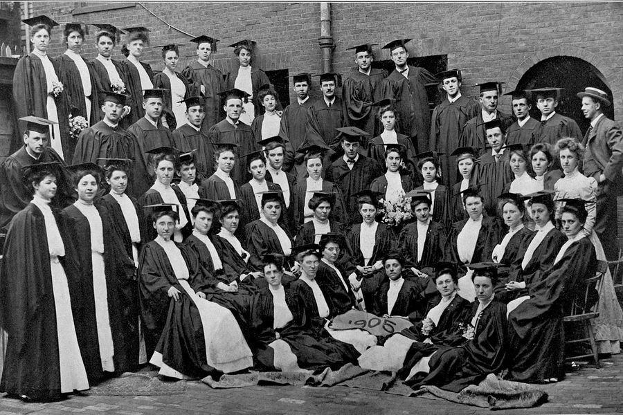 Photo: Black and white photo of a group of white men wearing graduation caps and gowns, and white women wearing white dresses and graduation caps and gowns, as they pose for a large group photo.