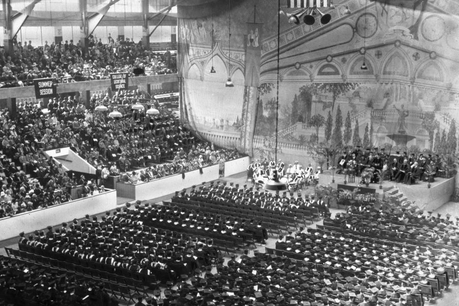 Photo: Black and white photo of 1927 Commencement. Boston Arena filed with students sitting in chairs in the middle, the diagonally diagonally seats are filled. The stage has a gigantic tapestry draped to make it appear the background is a courtyard.