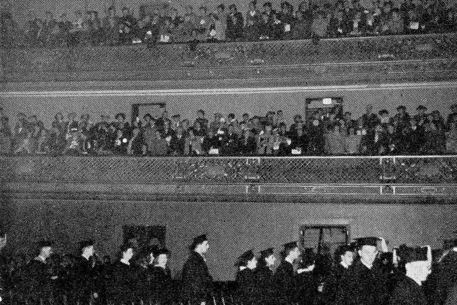 Photo: A very fuzzy black and white photo of the senior class filing into Symphony Hall for the 1945 Commencement. The seniors, in the bottom portion of the photo, are in their cap and gown. The top portion, with two rows of balcony space, are filled with the family members of those graduating.