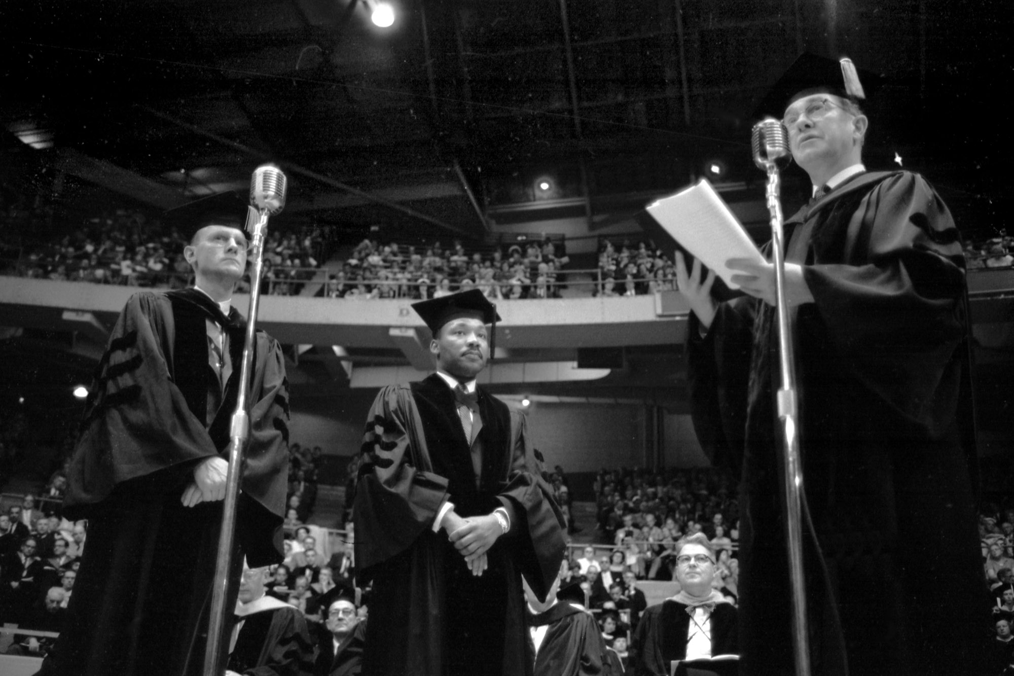 Photo: A black and white photo, a downward angle showing two mics in the foreground. Walter Muelder, Martin Luther King, Jr., and Boston University President Harold Case (left to right) at 1959 Boston University Commencement in Boston Garden. The audience is behind them.
