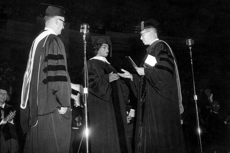 Photo: A black and white photo of three individuals in ceremonial garb. Two mics stand in the foreground. From left, a white man facing Singer, Marian Anderson, a Black woman, going to shake the hand of the white man, right, presenting here with her honorary degree.