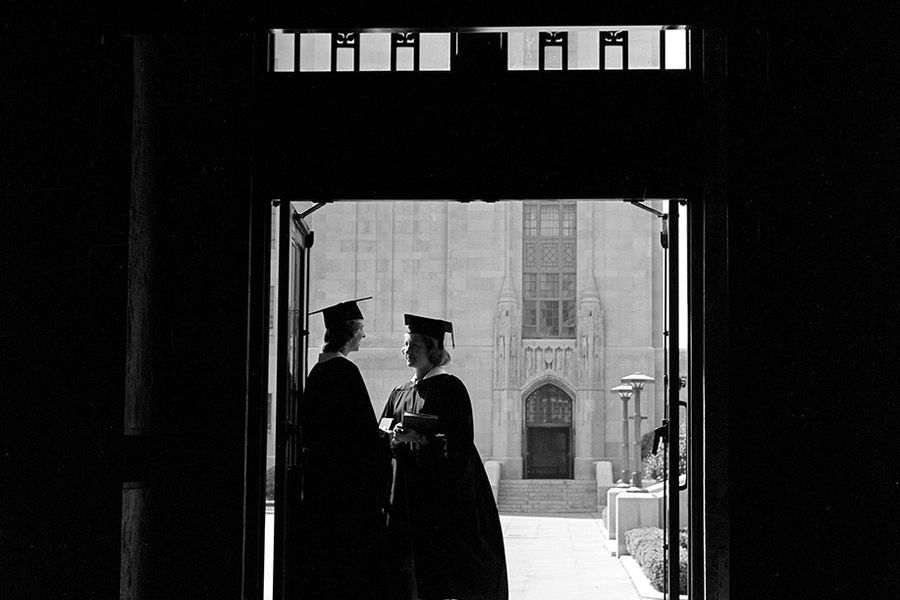 Photo: A black and white photo of two woman in the arched doorway of the CAS building, dawned in their cap and gown. They face each other, immersed in conversation.