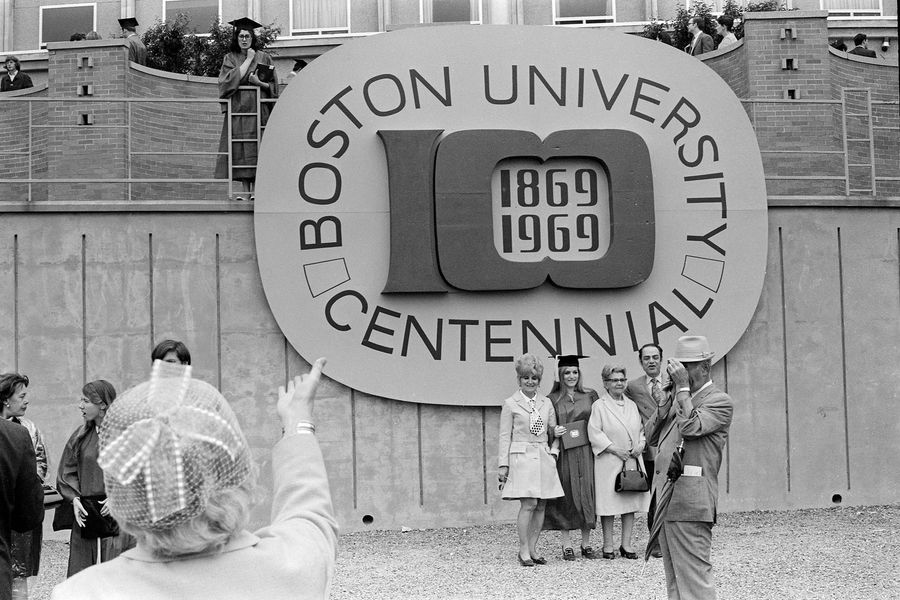 Photo: A black and white photo of people from commencement, four taking photos in front of Boston University's Centennial signage, one (a man in a full suit) taking a photo of something off screen, another (the woman in a foreground with a tulle hat). There are various other participants scattered in the photo.