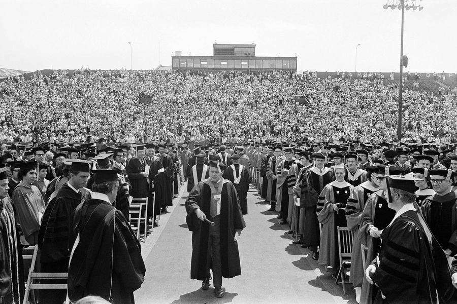 Photo: A black and white photo of the processional for the 1972 commencement. The faculty walk down the aisle with their ceremonial garb. Flanking the left and right of the photo are the graduates in their cap and gown. The audience peers on in the background.