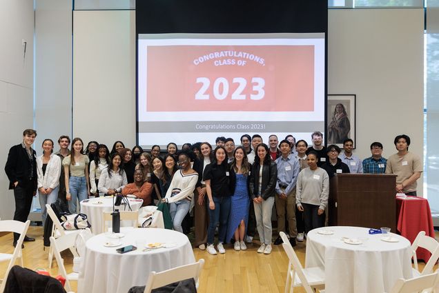 Photo: A large group of young adults dressed in business casual clothes smiles and pose together. A large projection shown behind them reads "Congratulations Class of 2023".