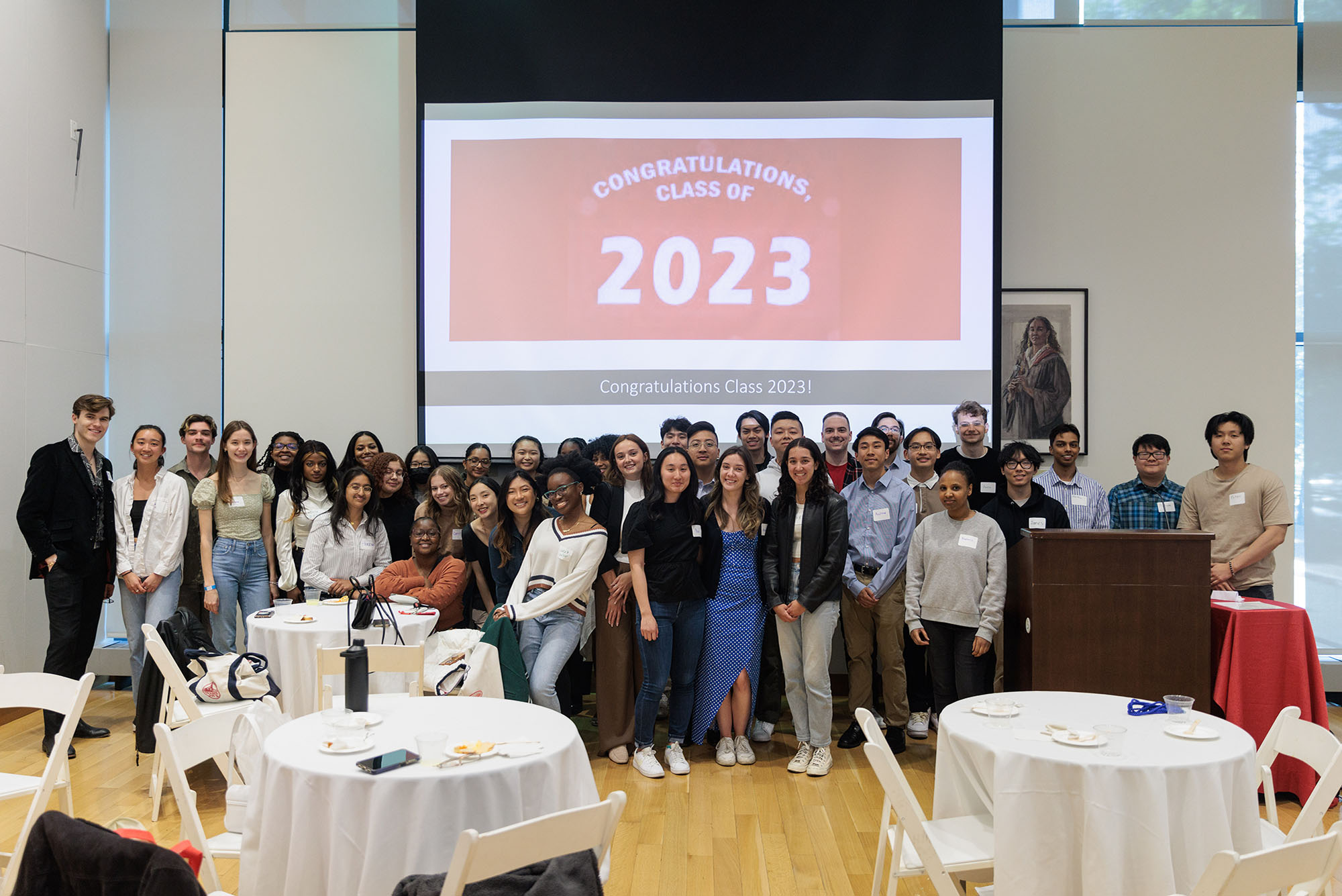 Photo: A large group of young adults dressed in business casual clothes smiles and pose together. A large projection shown behind them reads "Congratulations Class of 2023".
