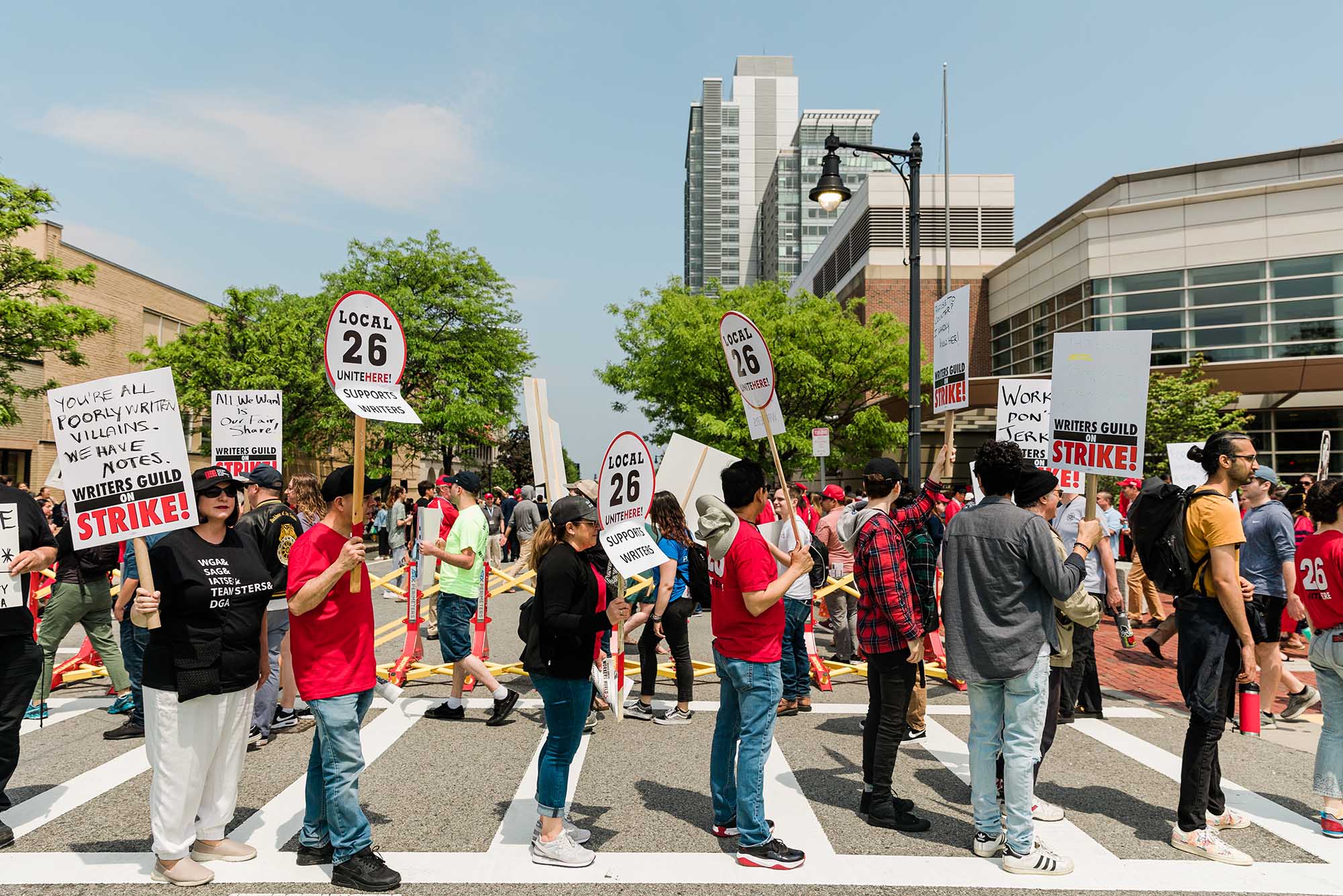 Photo: Two lines of protestors supporting the writer's strike stand outside Harry Agganis Way protesting the 2023 commencement speaker, David Zaslav. They hold up signs reading "SUPPORT WRITERS" and "YOU'RE POORLY WRITTEN VILLIANS, WE HAVE NOTES".