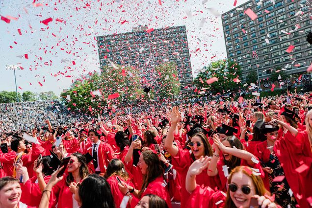 Photo: Graduates wearing red gowns and black graduation caps cheer and celebrate at the end of the 150th Boston University Commencement on Nickerson Field as red and white confetti falls from the air onto them.