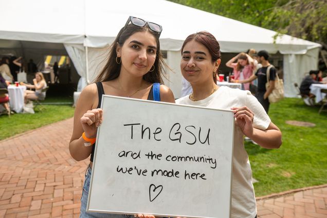 Photo: Two female students with long dark hair and one wearing glasses on their head, hold a sign that reads "The GSU and the Community we've made here"