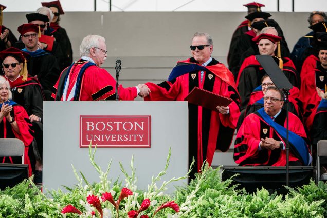 Photo: David Zaslav gives a speech at a podium at Boston University's 2023 commencement, while a sea of students look on