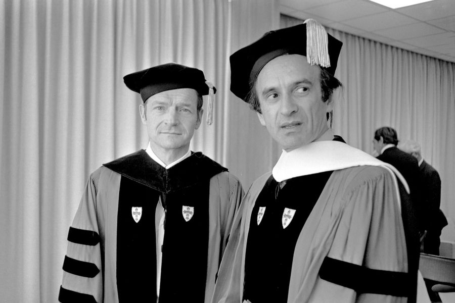 Photo: Two men, John Silber with Elie Wiesel, wearing formal university robes pose for a picture at a Boston University commencement. They are also wearing black caps, indicative of a high level of education. The photo is in black and white.