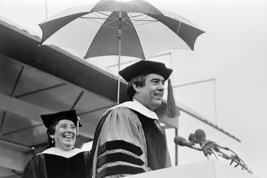 Photo: Jane Ahern, wearing a black cap and robes, holds an umbrella over the head of speaker Dan Rather, wearing a black cap and red robes, at Boston University's 1983 Commencement