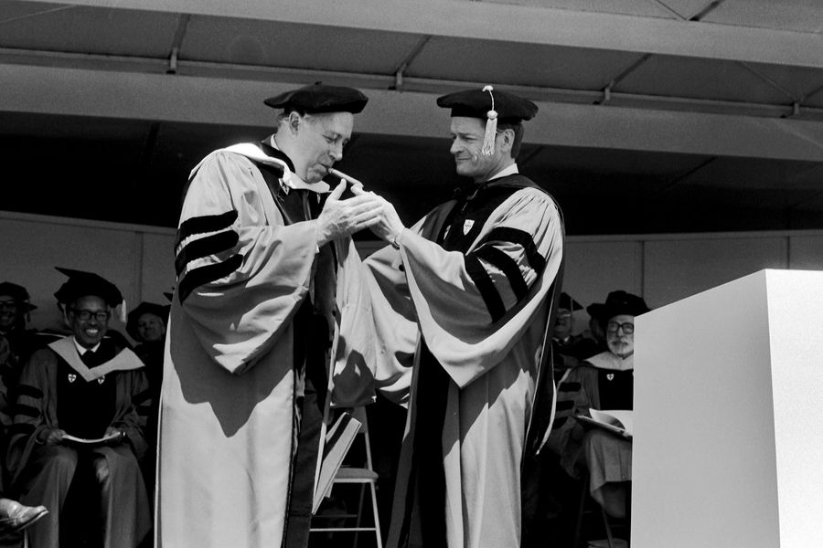 Photo: John Silber, in red robes with a black cap, lights Honorary Degree Recipient (similarly clothed) Red Auerbach's Cigar at the 1984 Commencement.
