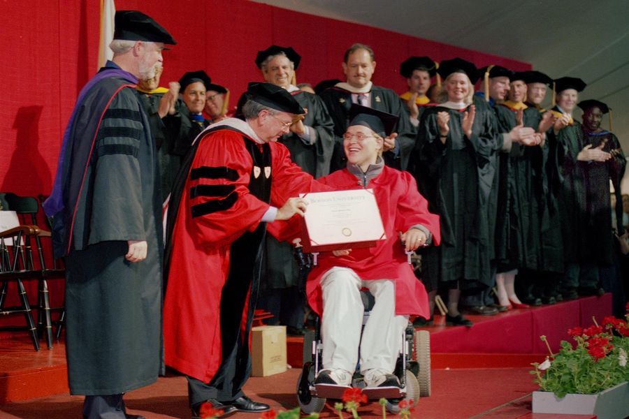 Photo: Travis Roy, a young white man in an electric wheelchair wearing a red graduation gown and black cap, smiles as he is handed a diploma on stage.
